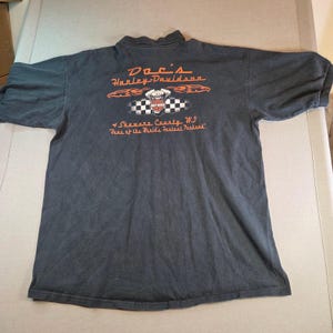 May include: Dark gray Harley-Davidson t-shirt with orange and white graphics. The back of the shirt features the text "Doc's Harley-Davidson" above a checkered flag design with a Harley-Davidson logo. Below the flag is the text "Shawano County, WI".