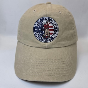 May include: A tan baseball cap featuring an embroidered patch with the text "2005 U.S. OPEN PINEHURST No.2". The patch depicts a golfer in a red vest and white hat. The cap has a curved brim and a button on top.