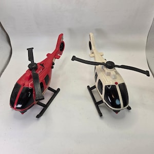 May include: Two toy helicopters. One is red with black accents and a black rotor. The other is off-white with black accents and a black rotor. Both have black landing skids and a black tail rotor.