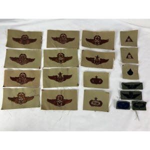 Op de afbeelding: Een verzameling vintage militaire patches, voornamelijk in beige en bruin. De patches tonen verschillende vleugel- en stermotieven, samen met andere insignes. Sommige patches hebben de tekst "US" en andere hebben een zwarte vorm.