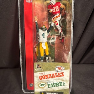 Puede incluir: Un paquete de plástico que contiene dos figuras de acción de la NFL. Las figuras son de Tony Gonzalez, un jugador de los Kansas City Chiefs, y Brett Favre, un jugador de los Green Bay Packers. Las figuras están en una pose dinámica, con Gonzalez atrapando un pase de Favre. El paquete es rojo, blanco y verde, con el logotipo de la NFL y los nombres de los jugadores y los logotipos de los equipos.