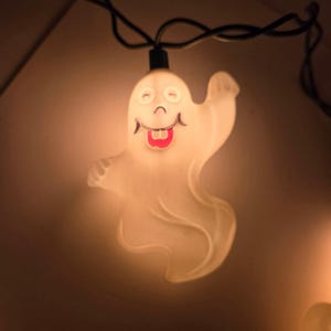 May include: A white, translucent, ghost-shaped light cover attached to a black electrical cord. The ghost features a smiling face with two small eyes, a curved mouth, and a bright red tongue, illuminated from within to emit a warm, soft glow.