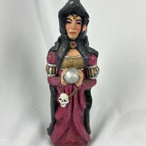 May include: A hand-painted figurine of a witch in a black pointed hat and cloak, holding a silver orb. The witch wears a red and gold dress with a skull pendant. The figurine is a decorative item.