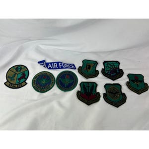 May include: A collection of embroidered Air Force patches in various shapes and designs. The patches feature green, blue, and red colors, with text such as "Air Force" and "Air Training Command". Some patches are circular, others are shield-shaped.