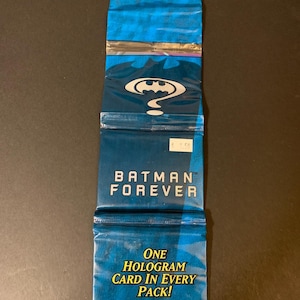 Puede incluir: Un paquete sellado de cartas coleccionables de Batman Forever en tonos azules. El paquete presenta el logotipo de Batman y el texto "BATMAN FOREVER". También indica "¡UNA CARTA HOLOGRÁFICA EN CADA PAQUETE!" y "17 CARTAS."