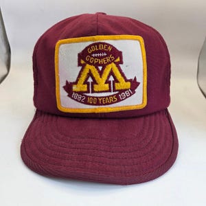 Puede incluir: Una gorra de béisbol de color granate con un parche rectangular en la parte delantera. El parche muestra una letra M amarilla, el texto Golden Gophers y las fechas 1882 y 1981 junto a la frase 100 Years. La gorra tiene una visera curva.