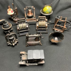 Puede incluir: Una colección de diez sacapuntas de metal en miniatura con varios diseños vintage, incluyendo dos cajas registradoras con la etiqueta 'CASH SALES', un globo terráqueo, una máquina de coser, un gramófono, una campana, una lámpara, una estufa, una bomba y un coche.