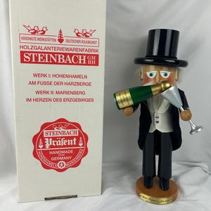 May include: A wooden Steinbach nutcracker figurine in a tuxedo holding a champagne bottle and a glass. The figurine has a black top hat and is standing on a wooden base. A box with German text is to the left.