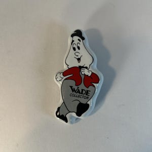 May include: A white ceramic figurine of a cartoon character wearing a red shirt, black pants, and a black hat. The character is walking and has a big smile. The figurine has the text "WADE COLLECTOR" on the front.