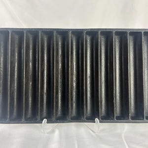 May include: A rectangular black cast iron corn stick pan featuring eleven parallel, elongated, semi-cylindrical molds. The pan has a flat handle on each of the two shorter sides, with a single circular hole centered in each handle for hanging.