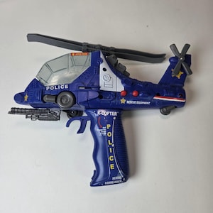 Könnte beinhalten: Ein blau-weißer Spielzeughelikopter mit dem Wort "POLICE" auf der Seite. Das Spielzeug hat einen Griff und einen Auslöser, auf dem die Wörter "K-COPTER" und "AERIAL COMMANDER" aufgedruckt sind. Das Spielzeug hat auch einen Propeller und eine kleine Pistole.