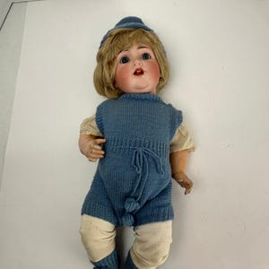 May include: A vintage doll with a painted face, blonde wig, and blue knit outfit. The doll wears a matching hat, romper, and booties. The doll's outfit includes cream-colored leg warmers. The doll is lying on a white surface.