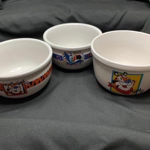 Może przedstawiać: Trzy białe ceramiczne miseczki na płatki śniadaniowe z kolorowymi grafikami postaci Kellogg's. Jedna miseczka przedstawia Tony'ego Tygrysa w pomarańczowe paski, druga Tukana Sama z kolorowymi kółkami, a trzecia Tony'ego Tygrysa w kwadracie.