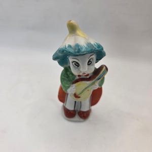 Puede incluir: Pequeña figura vintage de un músico. La figura lleva una chaqueta verde, una capa roja y un sombrero con detalles amarillos, blancos y azules. Sostiene un instrumento de cuerda amarillo y marrón. La figura tiene una cara blanca con ojos negros y labios rojos.