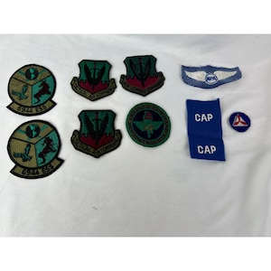 May include: A collection of military patches and insignia. The patches feature various designs, including eagles, wings, and text such as "Tactical Air Command" and "Air Combat Command." There are also blue fabric pieces with the word "CAP" and a small circular patch.