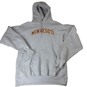 Peut inclure: Un sweat-shirt à capuche gris chiné à manches longues avec une poche kangourou sur le devant et un cordon de serrage. Le mot MINNESOTA est imprimé en arc de cercle sur la poitrine en lettres marron avec un fin contour jaune.