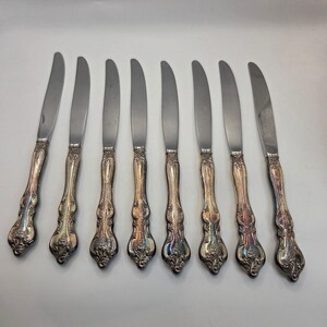 May include: A set of eight silver butter knives. Each knife features a stainless steel blade and an ornate, detailed handle with a floral design. The handles have a tarnished, antique appearance. The knives are arranged on a white surface.