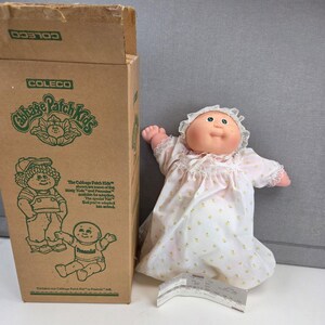 May include: A Cabbage Patch Kids doll with a soft vinyl head and fabric body, wearing a white nightgown with a yellow floral pattern and a matching bonnet. The doll is positioned next to its original brown cardboard box with green text.