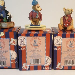 May include: Three boxed figurines, each with a different character. The first box features a jester figurine with a red and blue costume, sitting on a book. The second box features a monkey figurine with a blue hat and a red and blue costume. The third box features a teddy bear figurine with a red and white vest. Each box has a blue and red striped design with the character's name and a description.