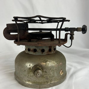 Könnte beinhalten: Ein tragbarer Vintage-Campingkocher mit einem verwitterten metallischen Brennstofftank als Basis und einem dunkel verrosteten Eisenbrenner. Das Gerät verfügt über einen kreisförmigen Rost, einen seitlichen Drehknopf und ein Brennstoffeinlassventil.
