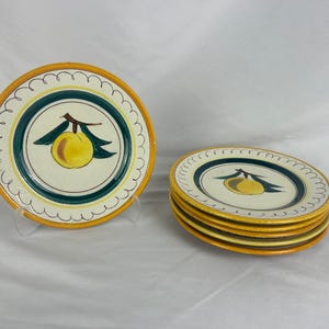May include: A set of decorative ceramic plates. Each plate features a yellow and green fruit design in the center, surrounded by a dark green and white border. The plates have a yellow scalloped edge. One plate is displayed upright, and the others are stacked.