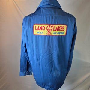 May include: A solid blue long-sleeve work jacket featuring a rectangular patch on the back. The patch has a yellow background with a red border and displays the red text LAND O'LAKES, the words MILK and ICE CREAM, and a central graphic logo.