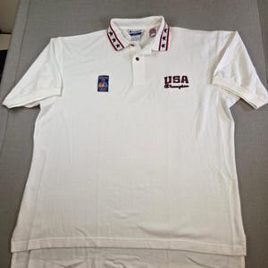 Puede incluir: Polo Champion blanco con cuello. El cuello tiene estrellas rojas, blancas y azules. Presenta el texto "USA Champion" y el logotipo de los Juegos Olímpicos de Atlanta 1996. Camisa de manga corta.