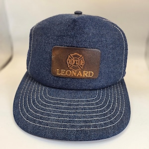 May include: A dark blue denim baseball cap featuring a rectangular brown leather patch on the front. The patch is embossed with a circular firefighter emblem containing the letters FD and the word LEONARD printed in capital letters below it.