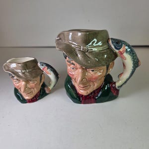 Könnte beinhalten: Zwei Keramikbecher in Form eines Männerkopfes mit Hut. Die Becher haben einen Fischgriff. Der größere Becher hat einen grün-roten Schal. Der kleinere Becher hat einen roten Schal.