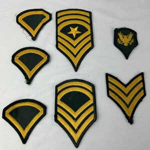 Peut inclure: Ensemble de patchs militaires vintage. Les patchs présentent un fond vert foncé avec des détails brodés or et noir. Divers motifs incluent des chevrons, des étoiles et un emblème d'aigle, représentant différents grades.