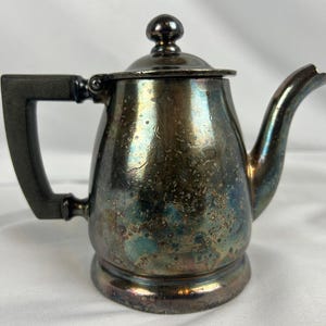 May include: A small, antique silver-colored teapot with a handle, spout, and lid. The teapot has a dark, tarnished appearance with iridescent hues of blue and green. The handle is a dark, rectangular shape. The teapot sits on a small, round base.