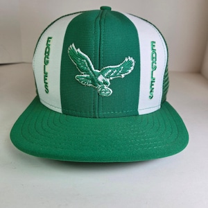 Puede incluir: Gorra de béisbol verde y blanca con el logotipo de los Philadelphia Eagles. La gorra tiene una visera plana y una parte trasera de malla. El nombre del equipo "EAGLES" está bordado en el lateral en verde. La parte delantera de la gorra muestra un águila verde.