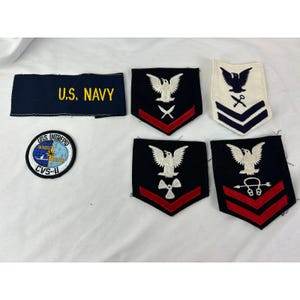 以下が含まれることがあります： アメリカ海軍のパッチと記章のコレクション。金色の文字で「U.S. NAVY」と書かれた青いパッチ、USS INTREPID CVS-11と書かれた円形パッチ、ワシとシェブロンが付いたさまざまな階級記章が含まれています。