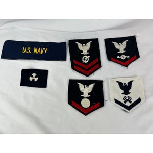 May include: A collection of U.S. Navy patches and insignia. Includes a dark blue patch with "U.S. NAVY" in gold lettering, along with several patches featuring eagles, chevrons, and other symbols. Some patches have red accents.