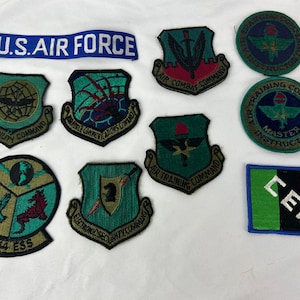 Op de afbeelding: Een verzameling geborduurde patches, waaronder een blauwe banner met "U.S. AIR FORCE" in witte letters. Verschillende patches tonen militaire insignes, schilden en teksten zoals "AIR COMBAT COMMAND" en "AIR TRAINING COMMAND". Kleuren zijn groen, rood en goud.