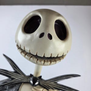 Peut inclure: Une figurine représentant une tête de crâne ronde blanc cassé avec de grandes orbites sombres et creuses ainsi qu'une bouche cousue. Sous la tête, un nœud papillon noir en forme de chauve-souris est fixé à un col de costume rayé sombre.