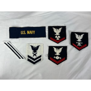 以下が含まれることがあります： アメリカ海軍のパッチと記章のコレクション。金色の文字で「U.S. NAVY」と書かれた濃い青色の長方形のパッチ、シェブロン、黒、白、赤のワシとシンボルが描かれたさまざまなパッチが含まれています。