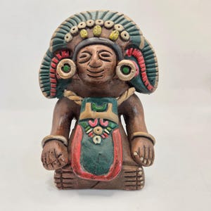 May include: A brown ceramic figurine of a seated figure with a detailed headdress and decorative chest piece. The figure has a serene expression and is adorned with green, red, and gold accents. The figurine is likely a representation of a historical or cultural figure.
