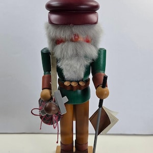 Peut inclure: Une figurine casse-noisette en bois portant un béret rouge foncé avec un pompon marron, une veste verte et un pantalon beige. La figurine a une barbe en fausse fourrure grise, porte une sacoche à carreaux avec une croix grise et une canne noire.
