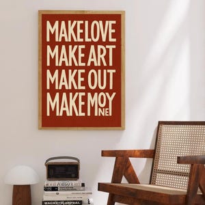 May include: A framed art print with the words "MAKE LOVE MAKE ART MAKE OUT MAKE MOY" in a bold, off-white font against a red background. The print is in a wooden frame. A wooden chair and books are in the foreground.