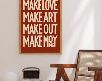 Puede incluir: Una impresión artística enmarcada con las palabras "MAKE LOVE MAKE ART MAKE OUT MAKE MOY" en una fuente en negrita, blanquecina sobre un fondo rojo. La impresión está en un marco de madera. Una silla de madera y libros están en primer plano.