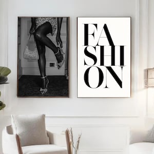 Puede incluir: Dos impresiones artísticas en blanco y negro enmarcadas. Una presenta a una persona ajustándose un zapato de tacón alto, sosteniendo un bolso. La otra impresión muestra la palabra "FASHION" en letras mayúsculas negras y en negrita sobre un fondo blanco. Las impresiones se muestran sobre una silla blanca.