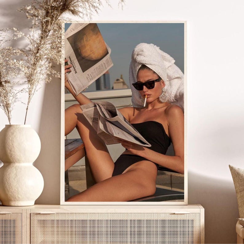 May include: A woman wearing a white towel on her head and a black swimsuit sits on a patio, reading a newspaper and smoking a cigarette.