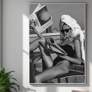 May include: A black and white photograph of a person wearing a white towel on their head and a black swimsuit. They are sitting on a chair and reading a newspaper while smoking a cigarette.