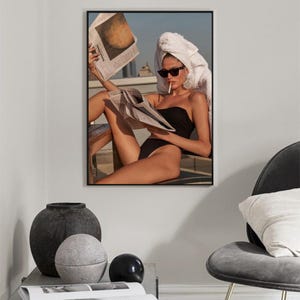 May include: A woman wearing a white towel on her head and sunglasses sits on a patio, reading a newspaper and smoking a cigarette. She is wearing a black swimsuit.