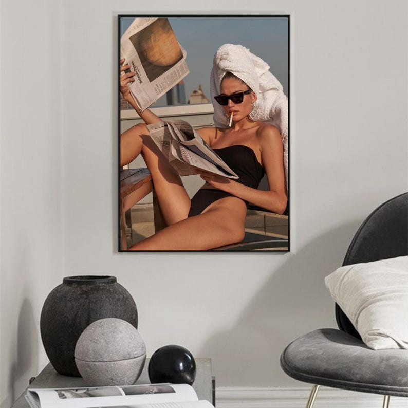 May include: A woman wearing a white towel on her head and sunglasses sits on a patio, reading a newspaper and smoking a cigarette. She is wearing a black swimsuit.