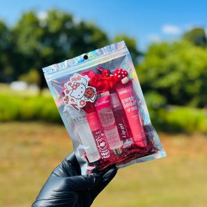 May include: A clear, holographic zip bag filled with lip glosses, lip scrub, and decorative items. The bag contains several pink lip gloss tubes, a red bow, a Hello Kitty sticker, and a mushroom-shaped charm. The background is a blurred outdoor scene.