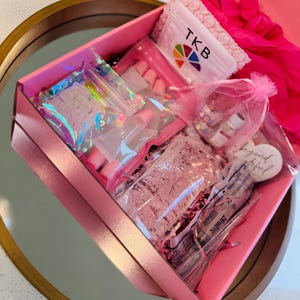 May include: A pink box filled with nail art supplies, including clear nail tips, glitter, and a packet with the letters "TKB" and a rainbow logo. The box is open, revealing various packaged items.