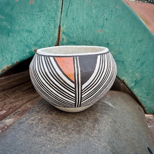 May include: A round, off-white ceramic bowl with a black, white, and orange geometric design. The bowl features vertical white lines against a black background, with an orange triangle shape. The bowl is sitting on a weathered surface.