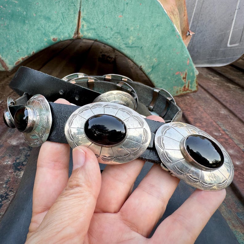 May include: Black leather belt with large, oval silver-toned buckles, each featuring a black stone center. The buckles have a decorative, sunburst-like pattern. The belt is held by a hand.